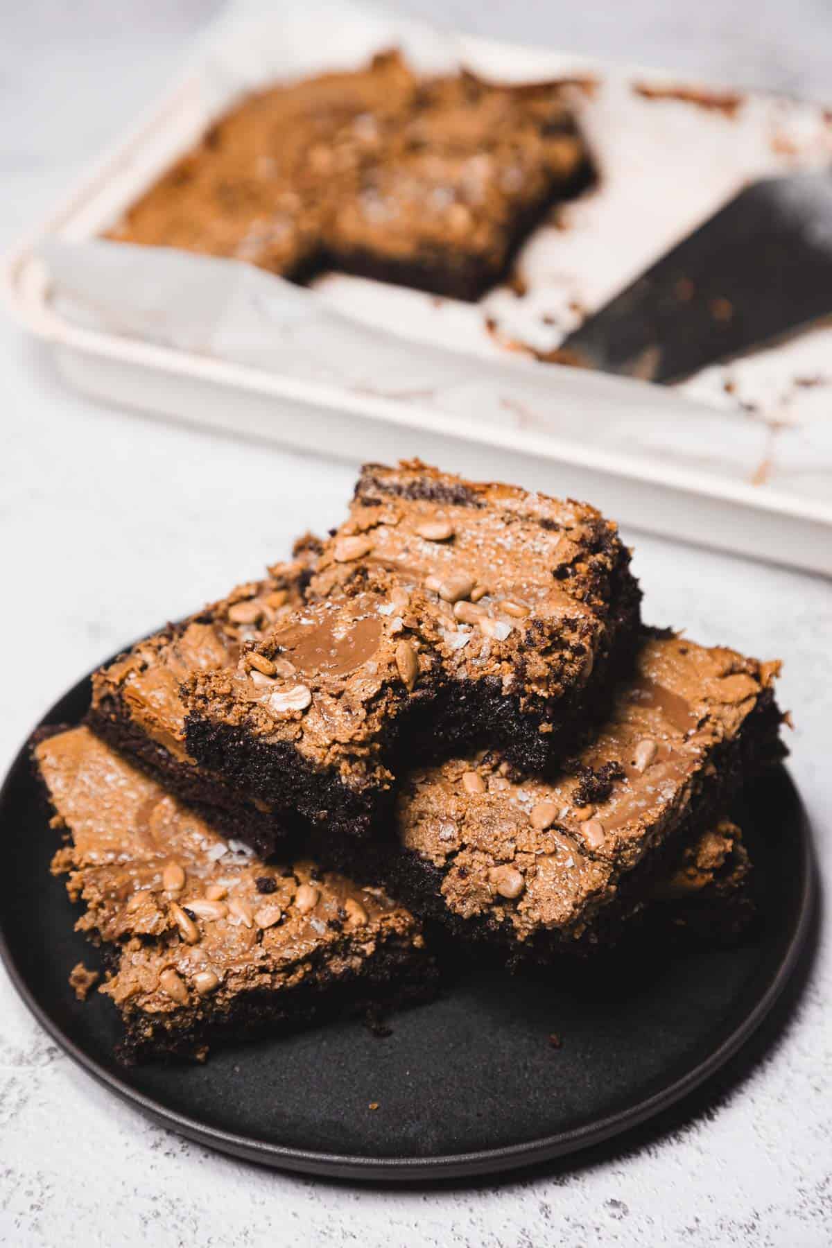 Three chocolate brownies stacked on a black plate, with more brownies in a tray in the background.