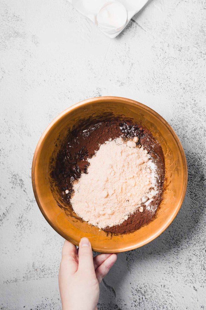 A hand holds a copper bowl with cocoa powder, flour, and other baking ingredients inside.