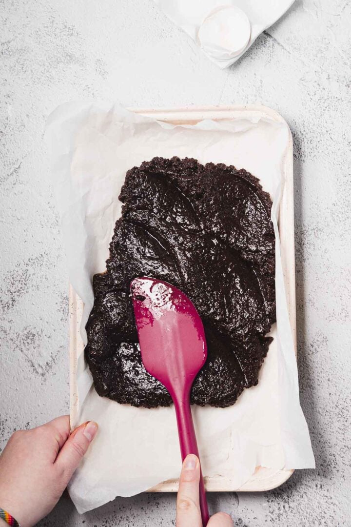 Hands spreading brownie batter in a parchment-lined pan with a pink spatula on a light surface.