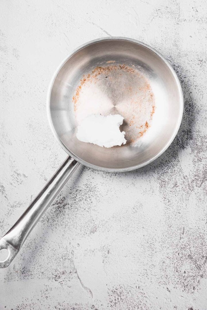 A stainless steel pan with a chunk of solid white coconut oil on a light textured surface.