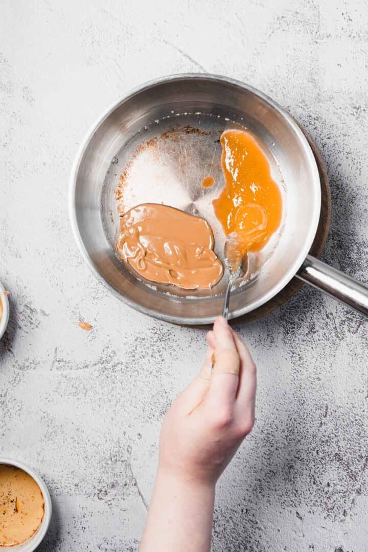A hand stirs melted caramel in a stainless steel pan on a light gray surface.