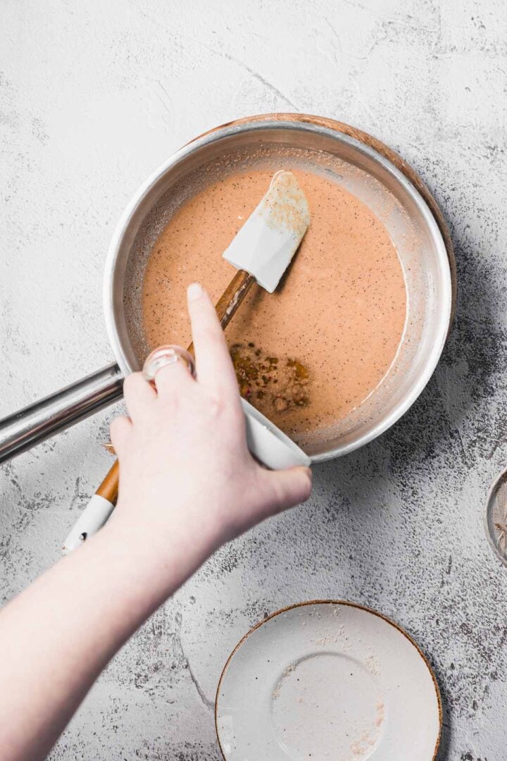 A hand stirs a light brown sauce in a saucepan with a spatula on a light gray textured surface.