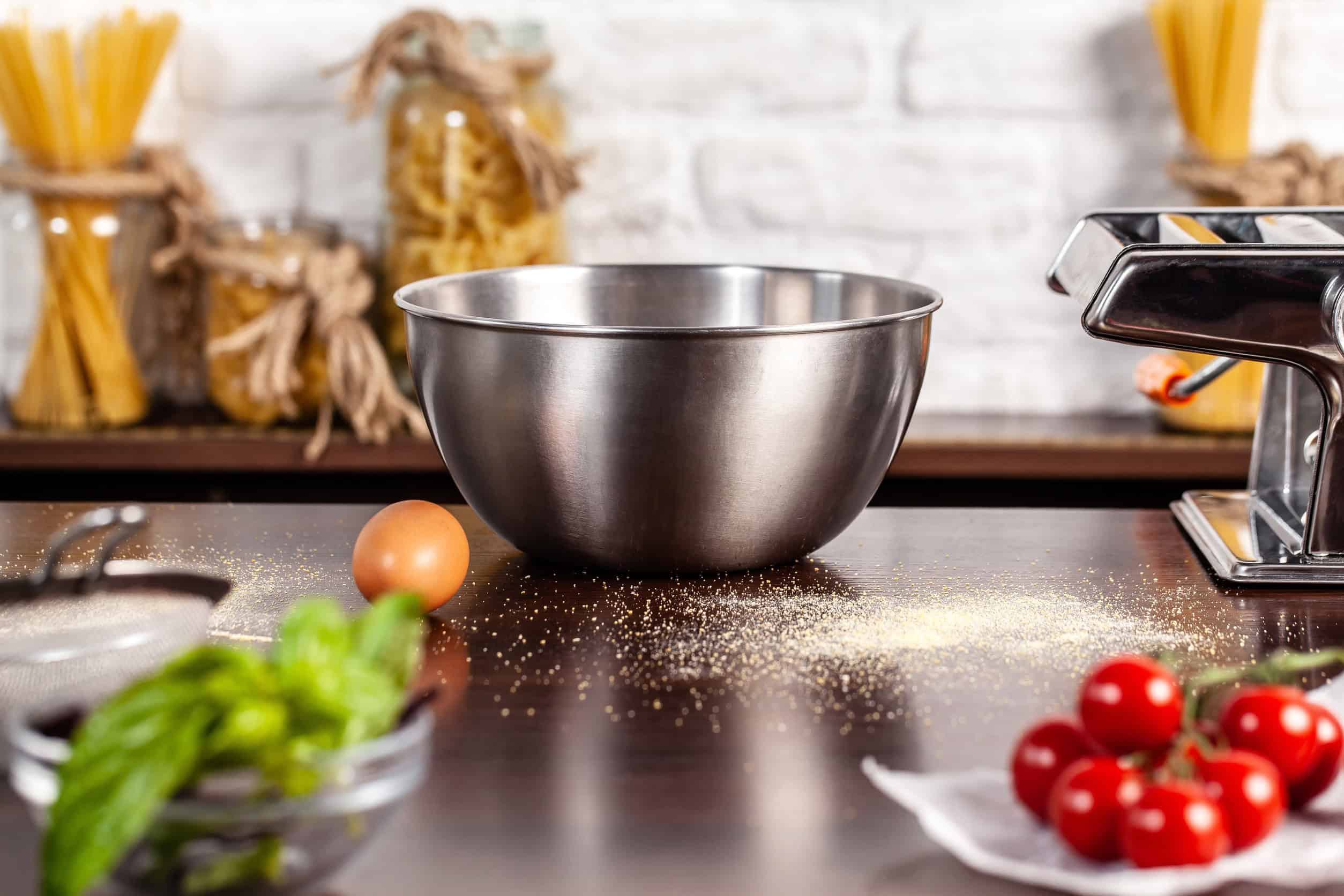 A metal mixing bowl on a kitchen counter with an egg, tomatoes, and pasta ingredients nearby—one of the essential cooking tools for beginners.