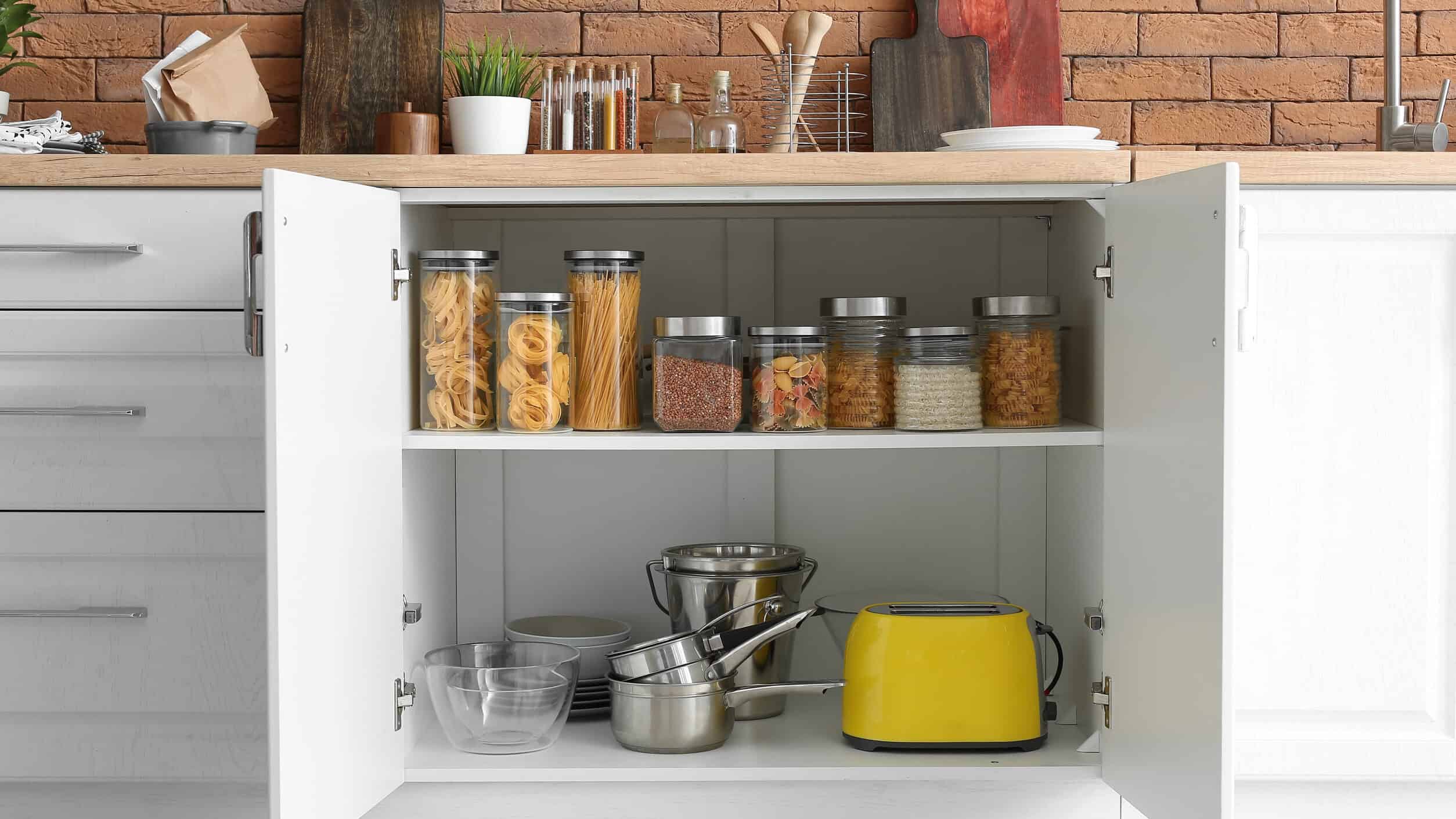 Open kitchen cabinet with jars of pasta and grains—perfect food storage gifts—on the top shelf, with cookware and a yellow toaster on the bottom shelf.