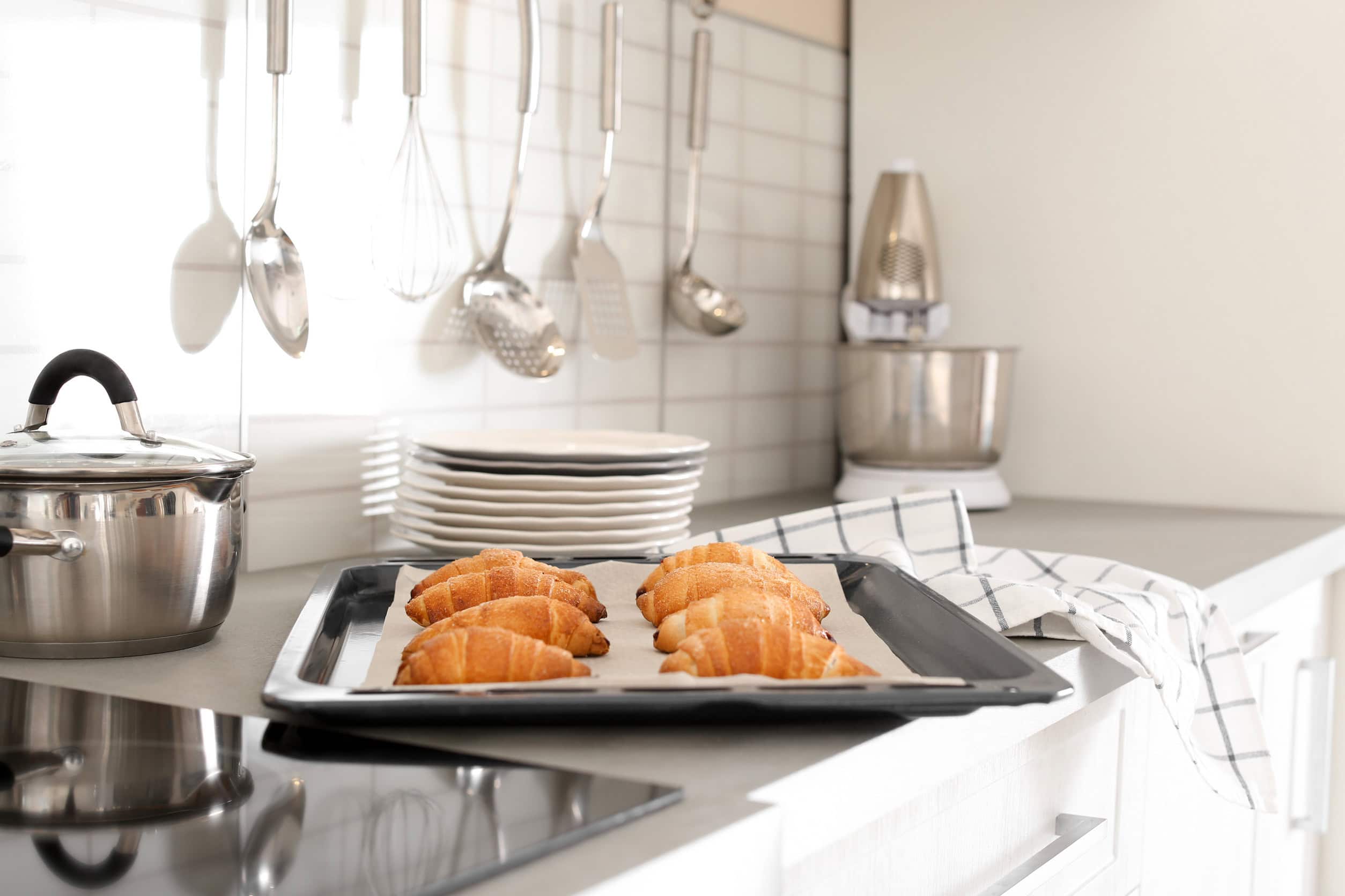 Freshly baked croissants on a tray in a modern kitchen, surrounded by essential cooking tools for beginners, with utensils and plates in the background.