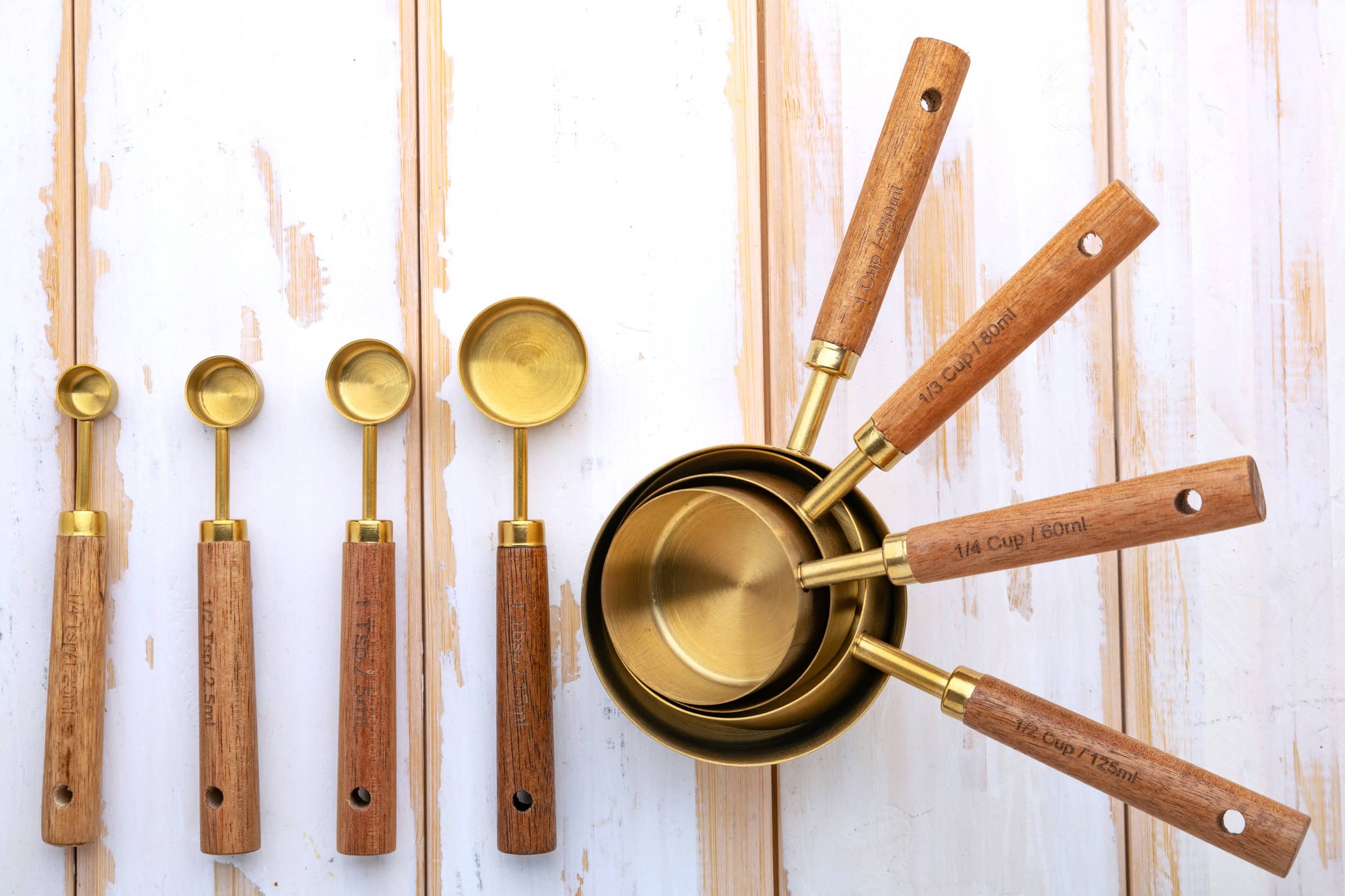 Gold measuring spoons and cups with wooden handles arranged on a white wooden surface—essential cooking tools for beginners.