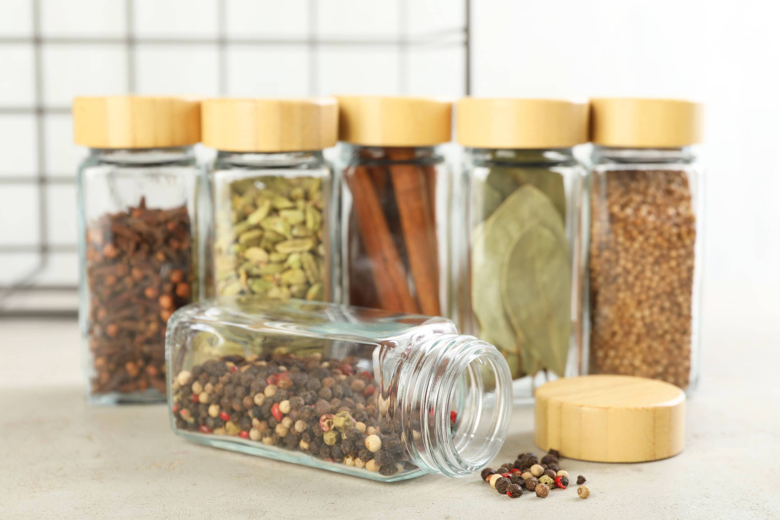 Six glass spice jars with wooden lids, perfect as food storage gifts, sit upright in the background while one lies down, filled with colorful mixed peppercorns.