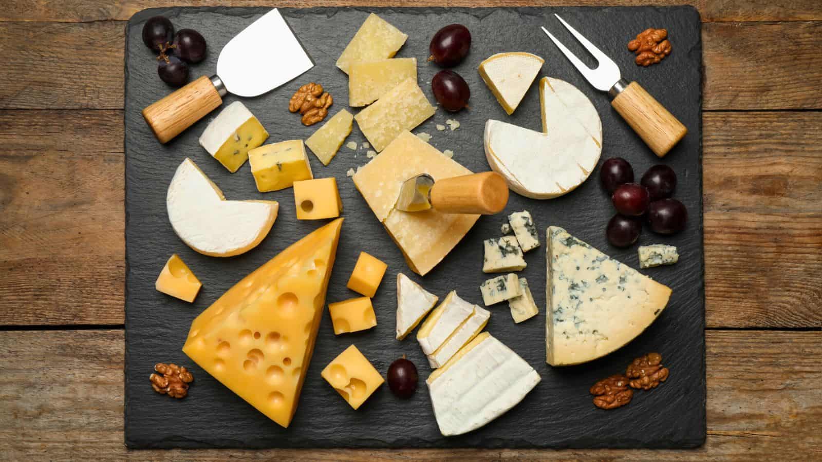 Assorted cheeses, grapes, and walnuts on a black slate platter with cheese knives—perfect gifts for cheese lovers—beautifully arranged on a wooden table.