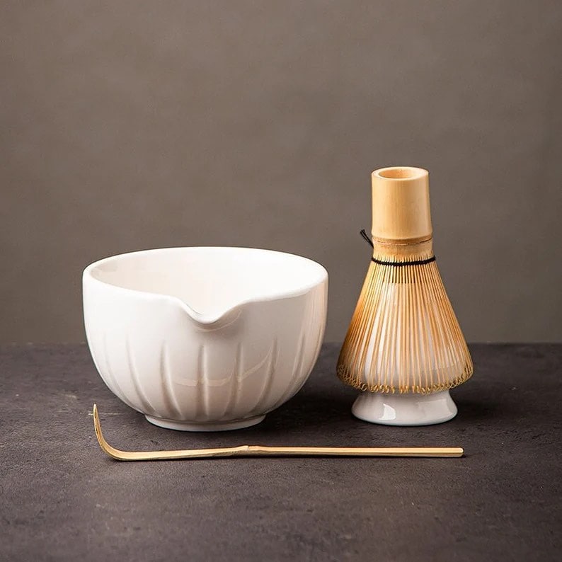 White matcha bowl, bamboo whisk, and metal tea scoop on a dark surface with a neutral background.