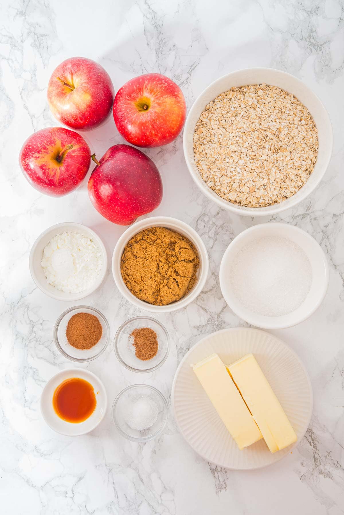 Top view of three apples, oats, butter sticks, sugars, spices, starch, and vanilla on a white marble surface.