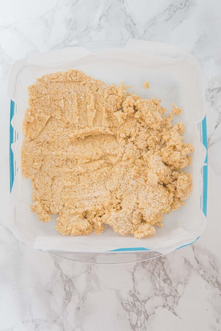 Oat mixture pressed into a parchment-lined square pan on a marble surface, ready for baking.