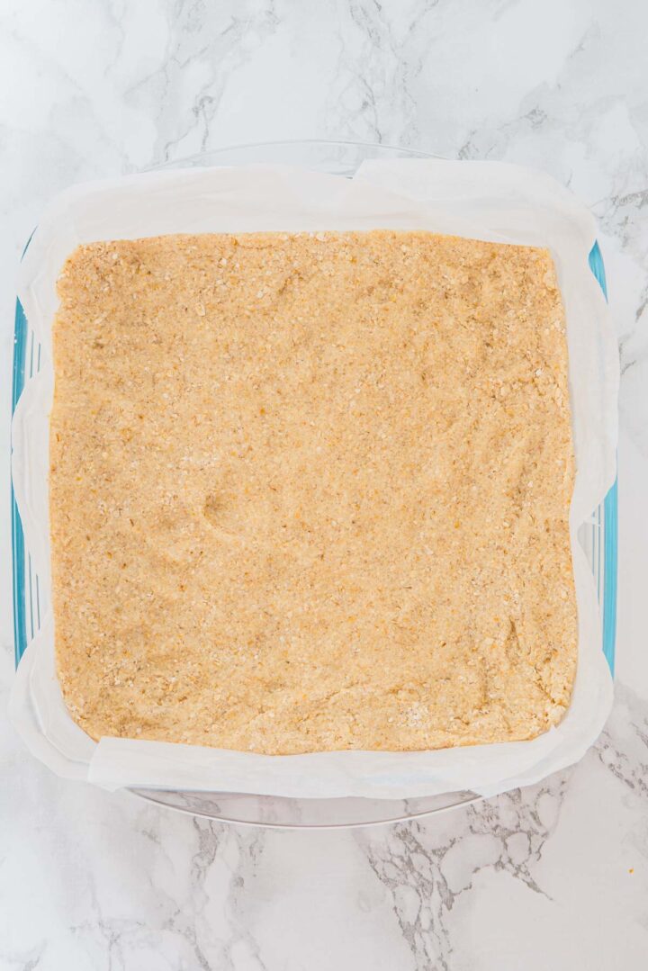 A parchment-lined baking dish filled with an even layer of unbaked crumbly crust on a marble surface.