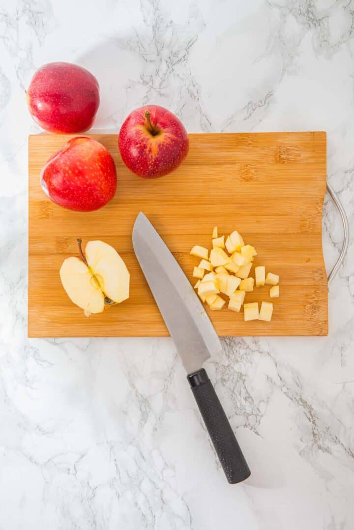 Three red apples, a knife, and diced apple pieces on a wooden cutting board on a marble surface.