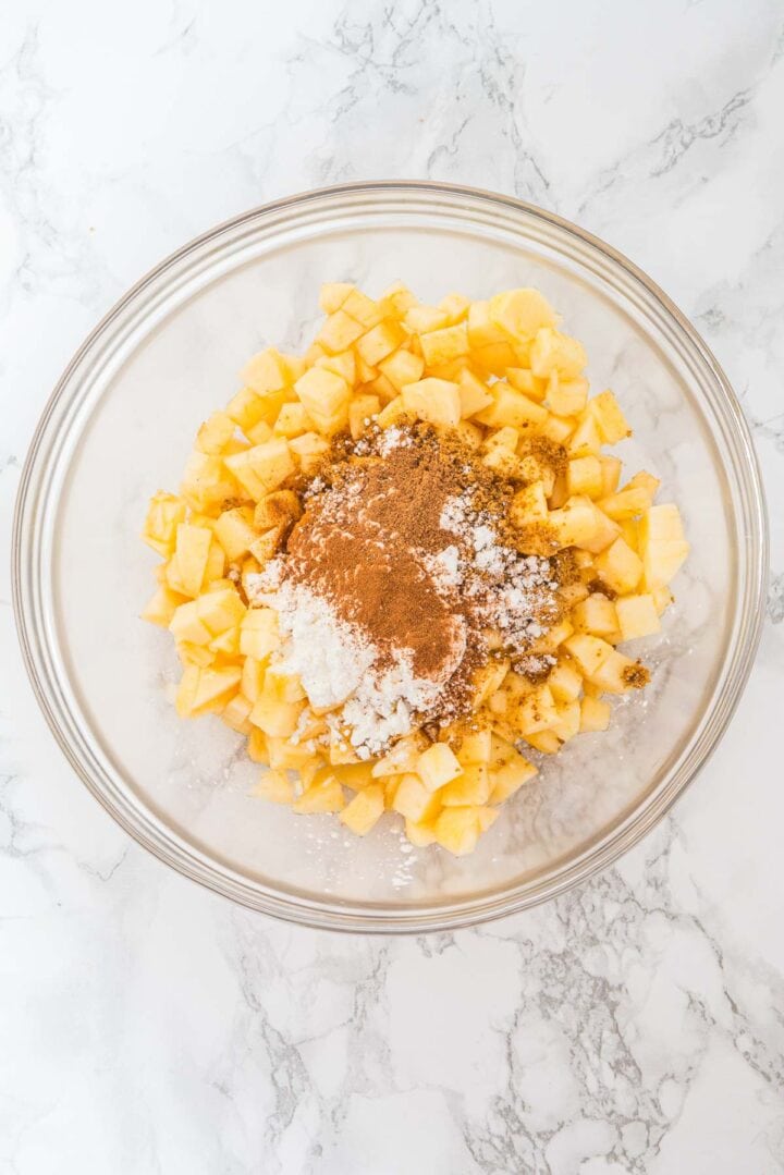 A glass bowl with chopped apples, cinnamon, sugar, and flour on a marble countertop.