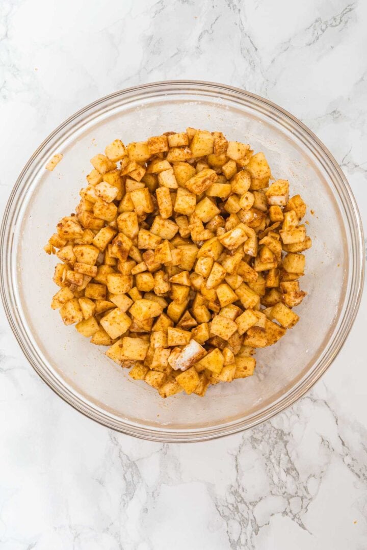 Diced apples coated with cinnamon and spices in a glass bowl on a marble surface.