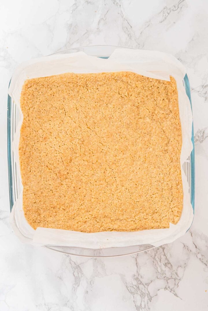 Square baking dish lined with parchment paper, filled with pressed oat mixture on a marble surface.