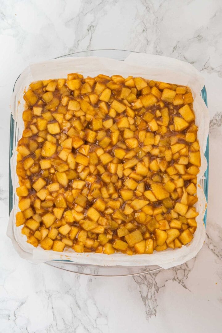 Baking dish lined with parchment paper filled with chopped apple pie filling on a marble surface.