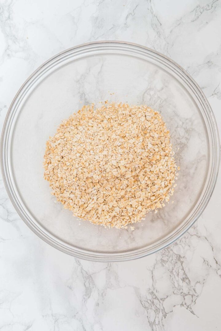 A glass bowl filled with dry oats on a white marble surface.
