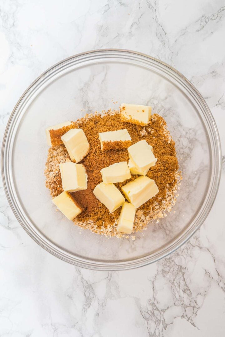 A glass bowl with oats, brown sugar, cinnamon, and chunks of butter on a marble surface.