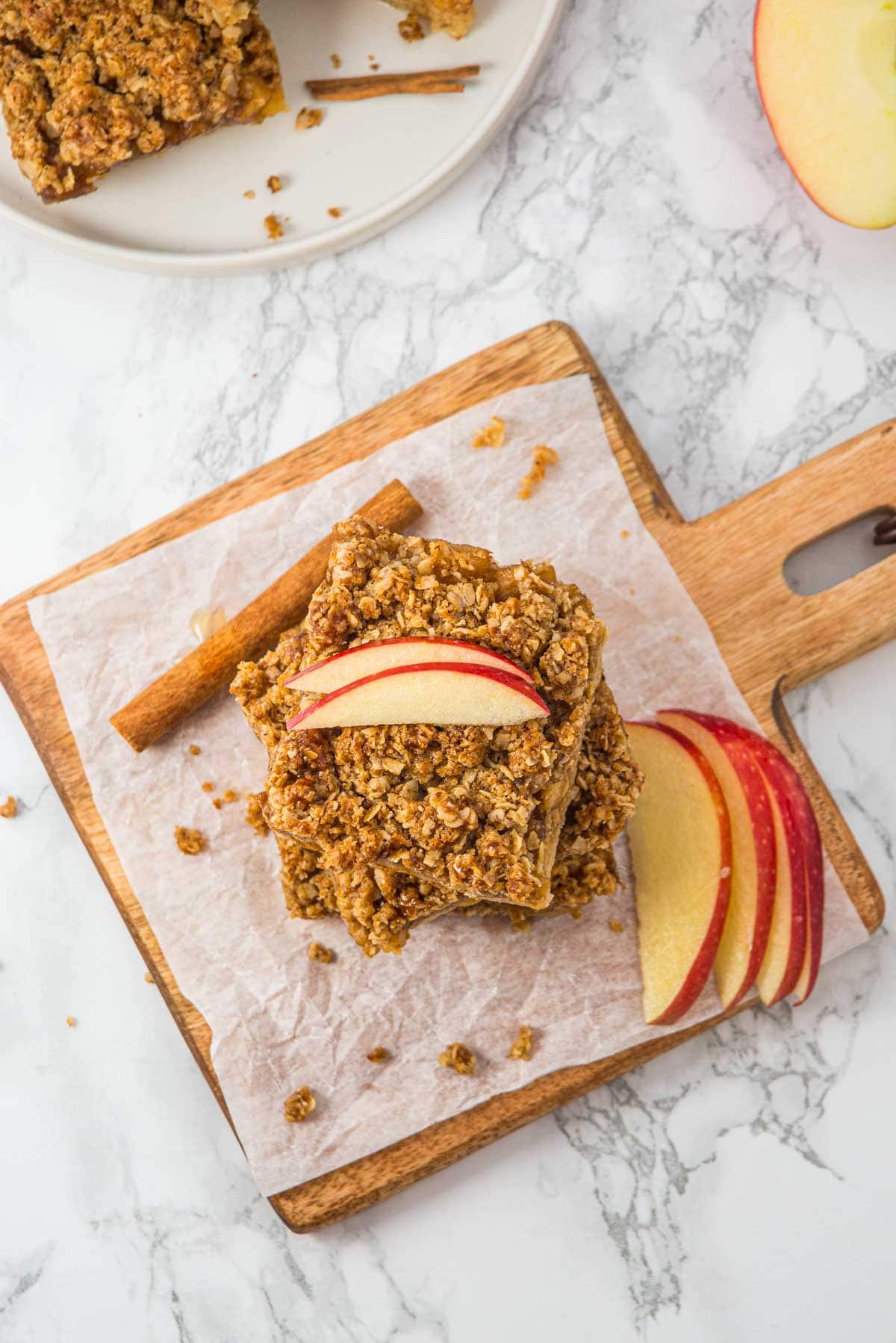 Oat crumble bars topped with apple slices on parchment paper, beside cinnamon sticks and sliced apples.