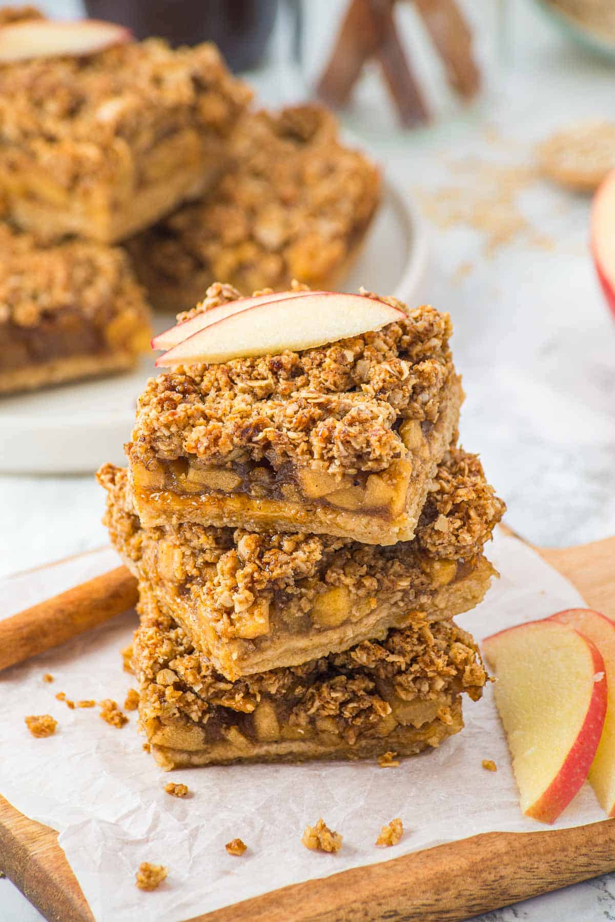 Three stacked apple crumble bars topped with a fresh apple slice on parchment paper, with more bars in the background.