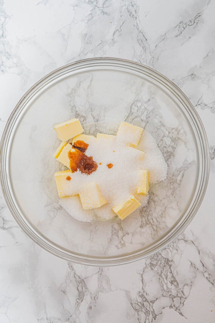 Glass bowl with cubes of butter, granulated sugar, and vanilla extract on a marble countertop.