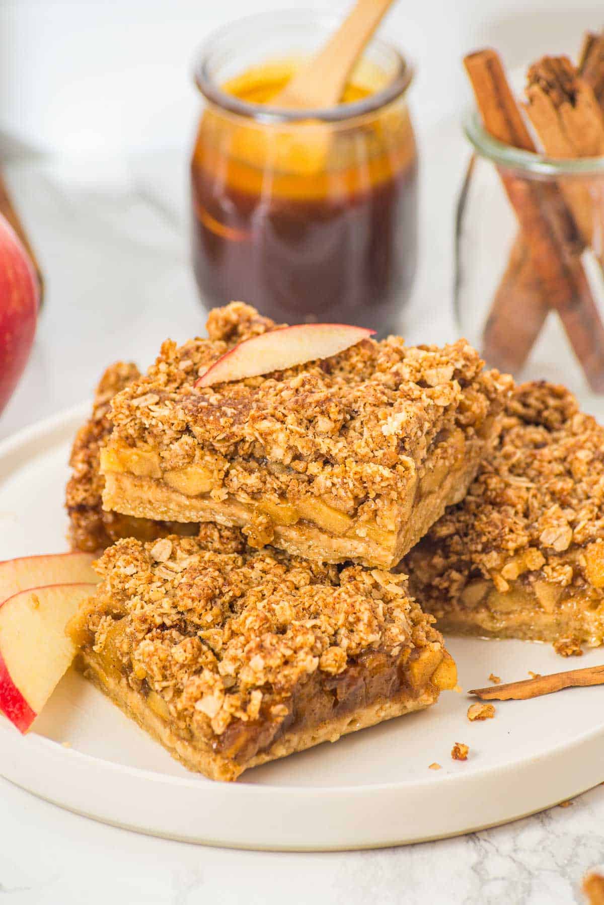 Three apple crumble bars on a plate, topped with oat streusel, with apple slices and cinnamon sticks nearby.