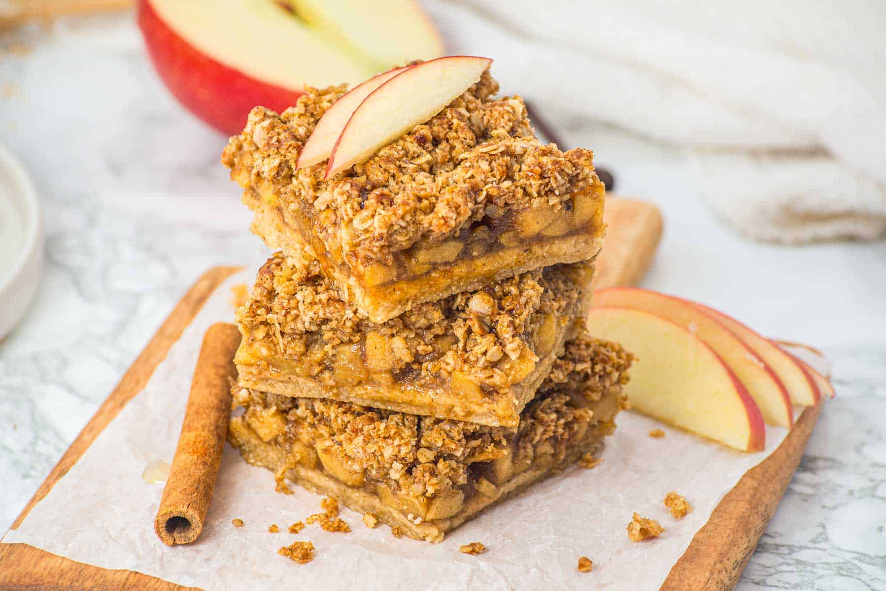 Three stacked apple crumble bars garnished with apple slices, on parchment paper with a cinnamon stick nearby.