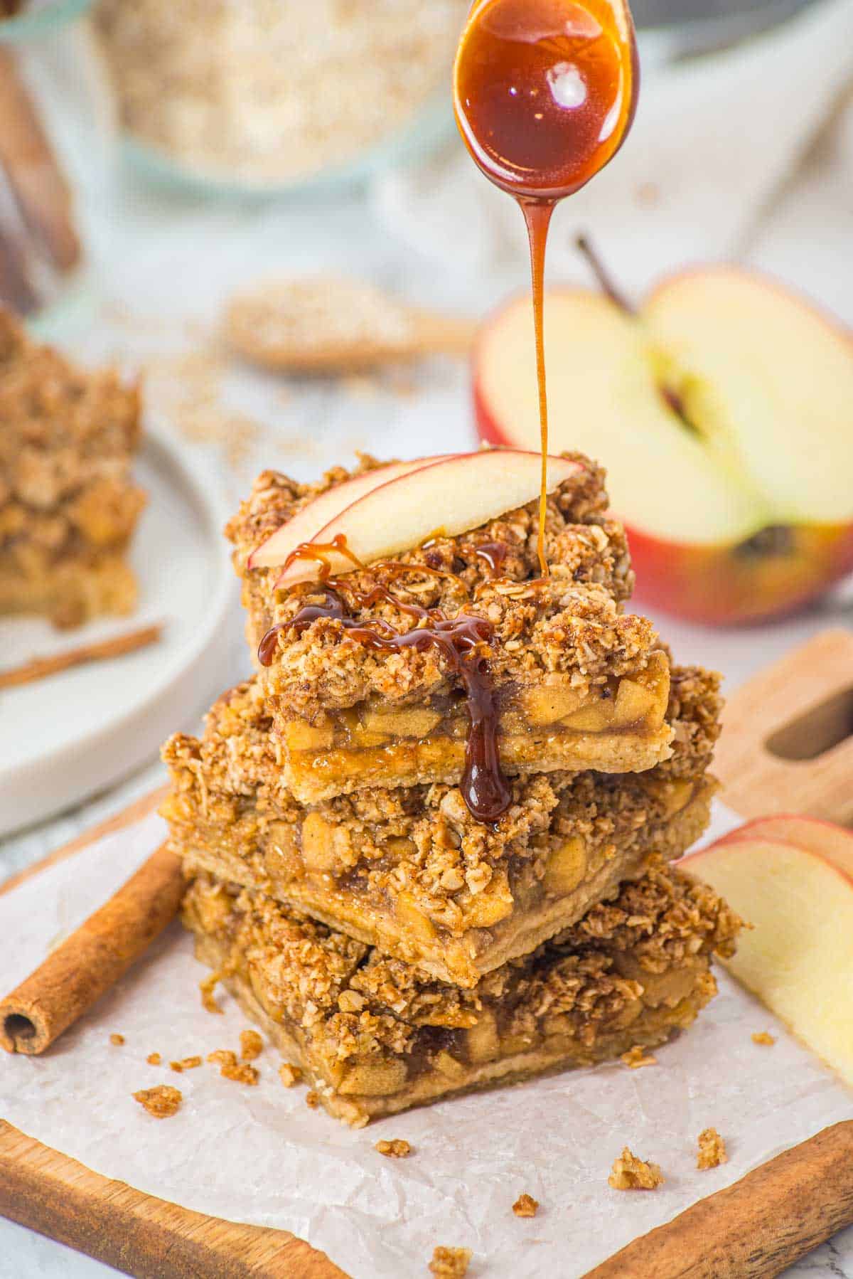 A stack of apple crumble bars is drizzled with caramel sauce, with an apple slice and cinnamon sticks nearby.