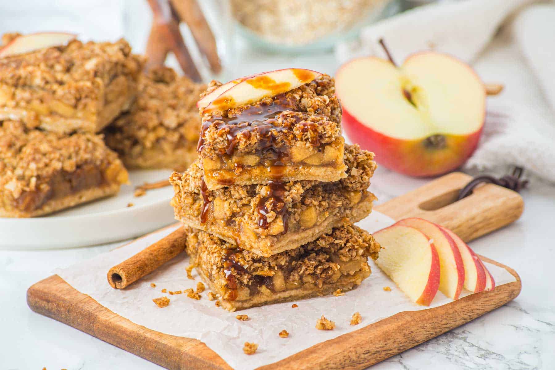 Three apple crumble bars stacked on a wooden board, topped with apple slices and caramel drizzle, with apples nearby.