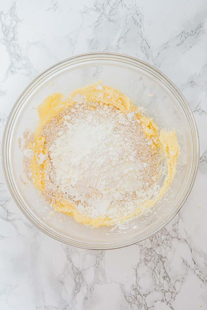 A glass bowl with flour and creamed butter mixture on a marble countertop, ready to be mixed.