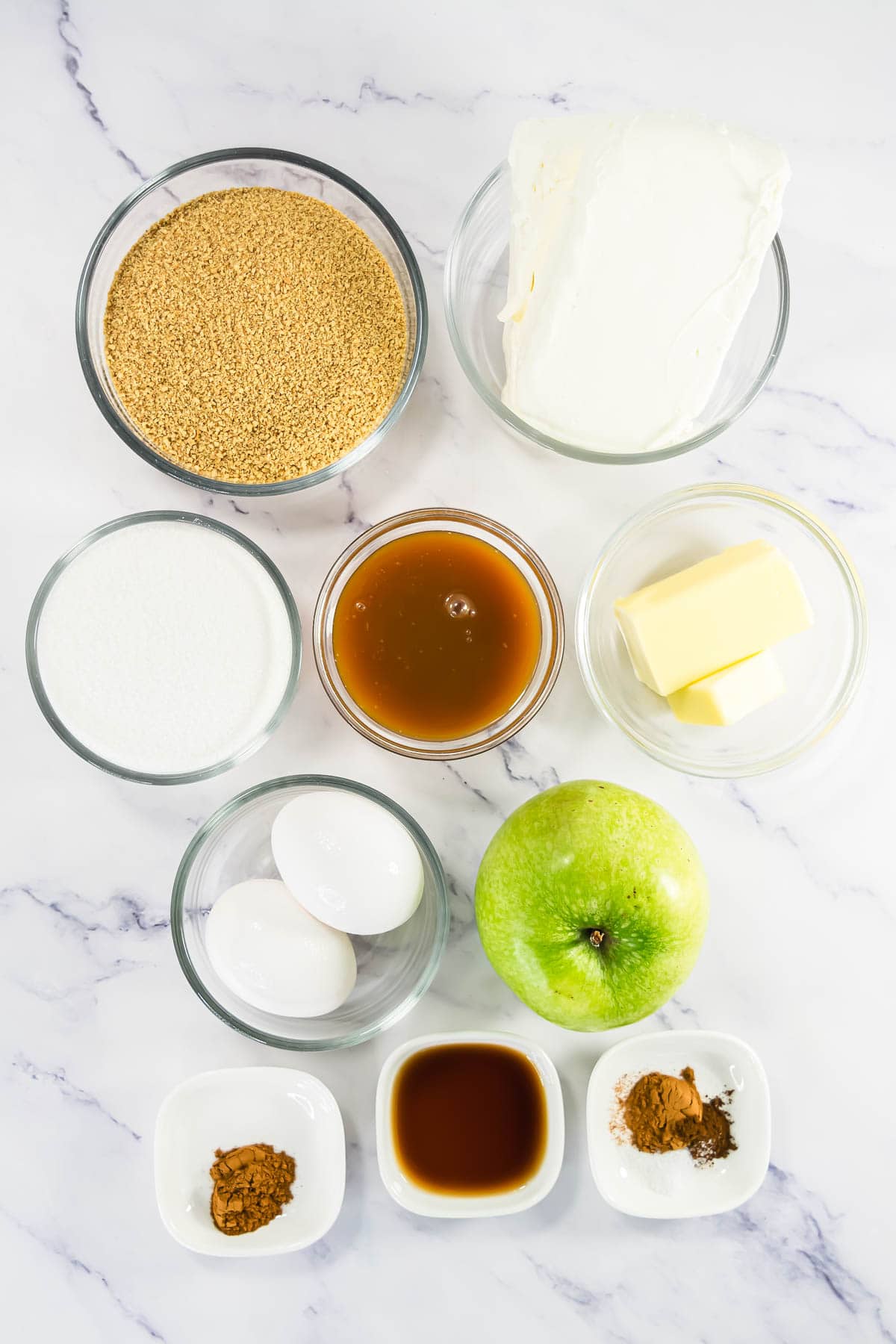 Ingredients in bowls: graham crumbs, cream cheese, sugar, caramel, butter, eggs, green apple, spices, vanilla.