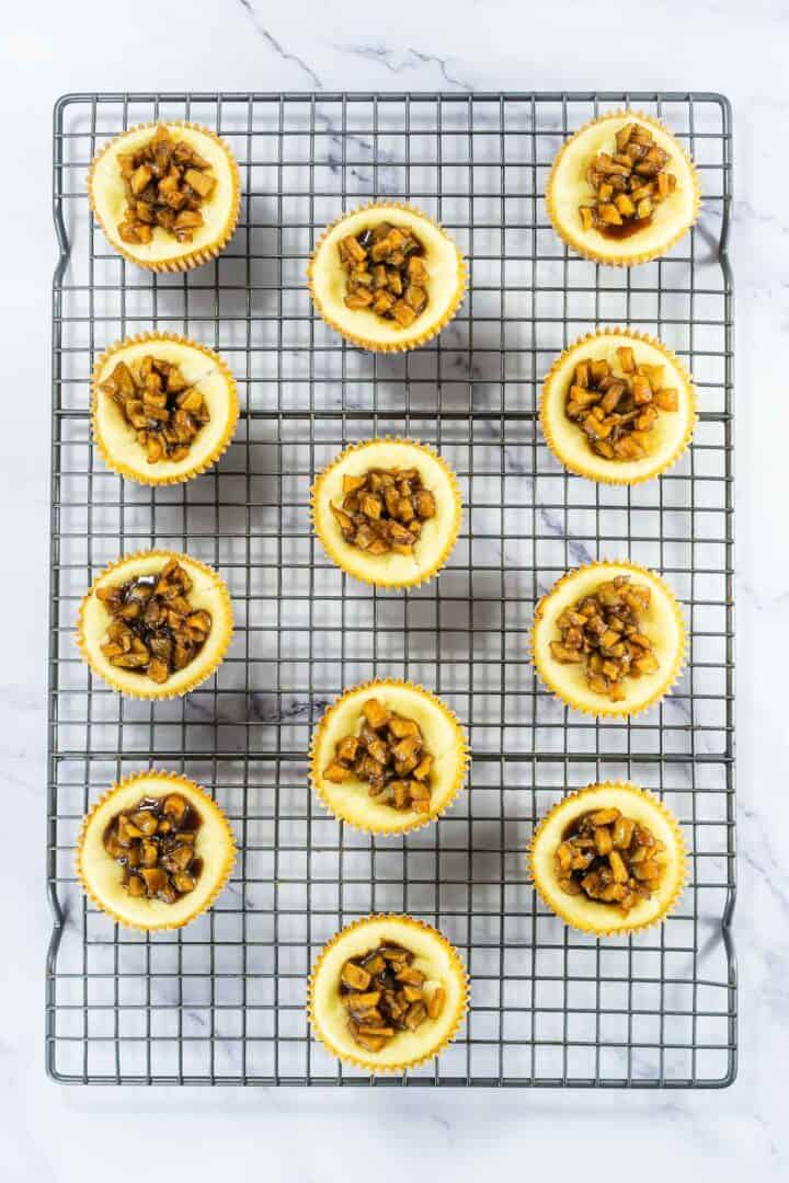 Mini cheesecakes topped with diced caramelized apples on a cooling rack, viewed from above.