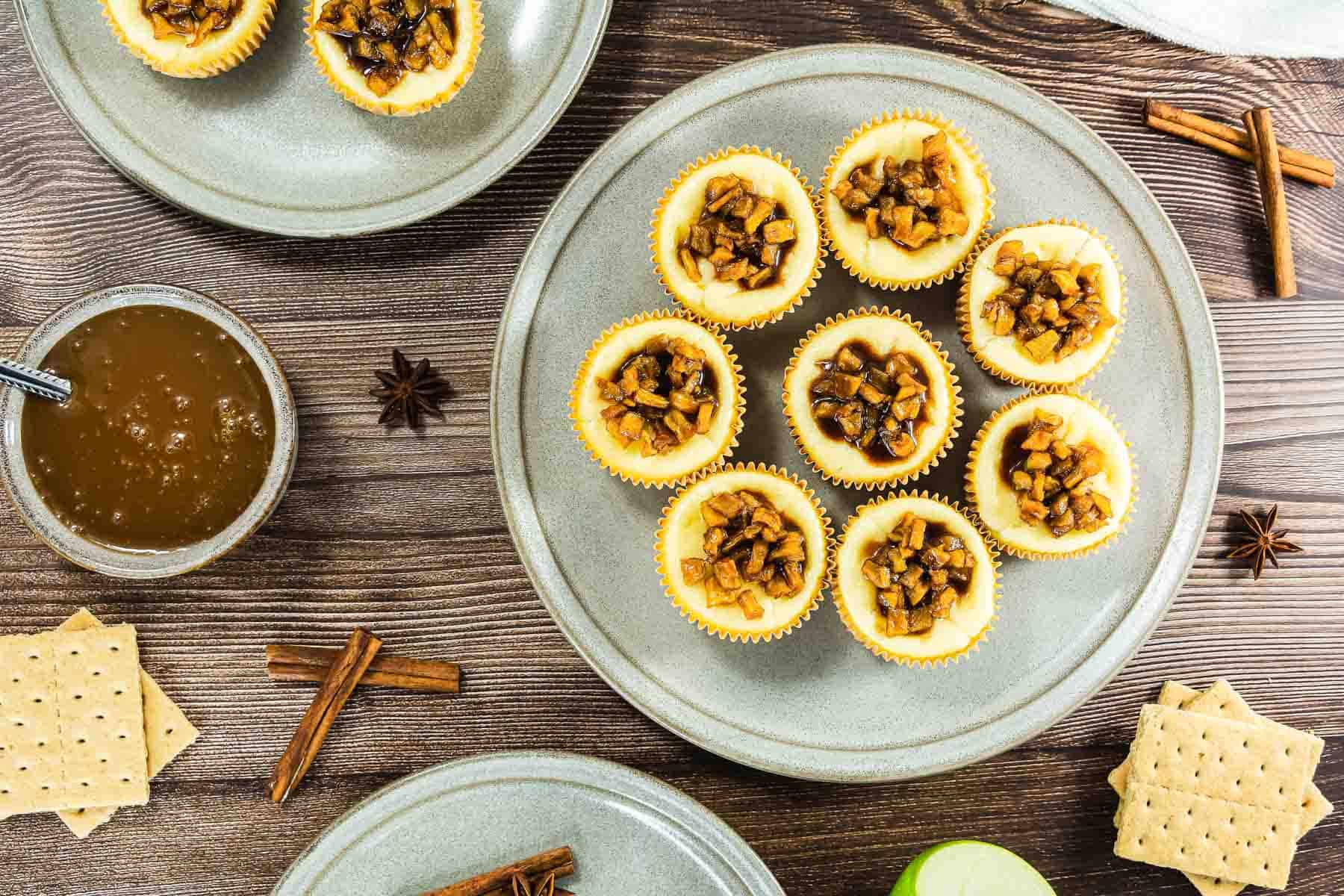 Mini cheesecakes topped with diced apples on a plate, surrounded by graham crackers and cinnamon sticks.