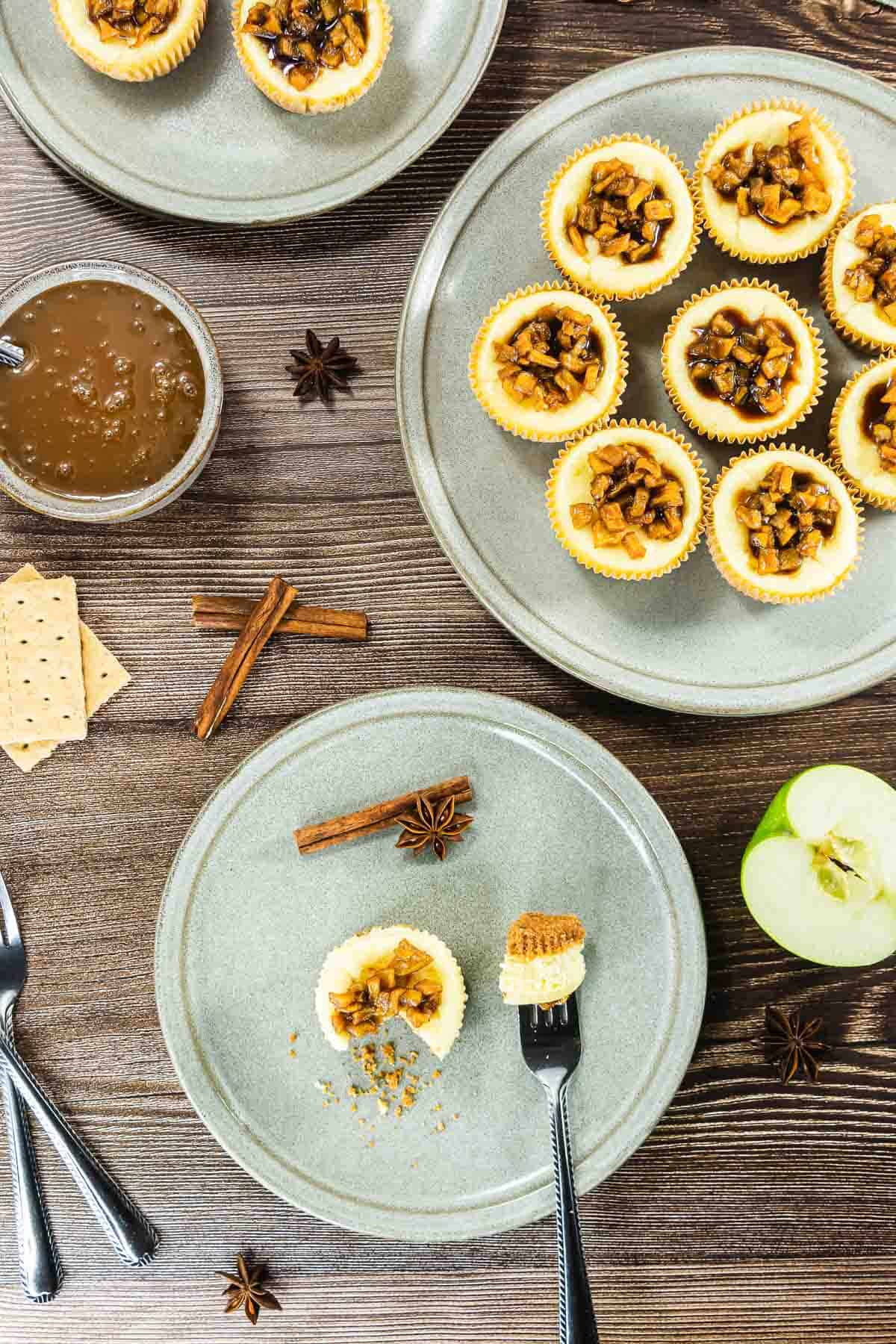 Mini apple cheesecakes on plates, with cinnamon sticks, anise, crackers, and a green apple on a wooden table.