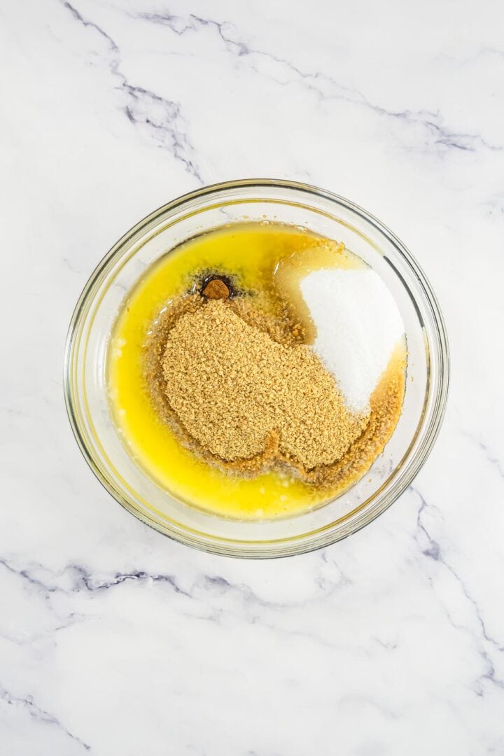 A glass bowl with melted butter, sugar, and graham cracker crumbs on a marble surface.