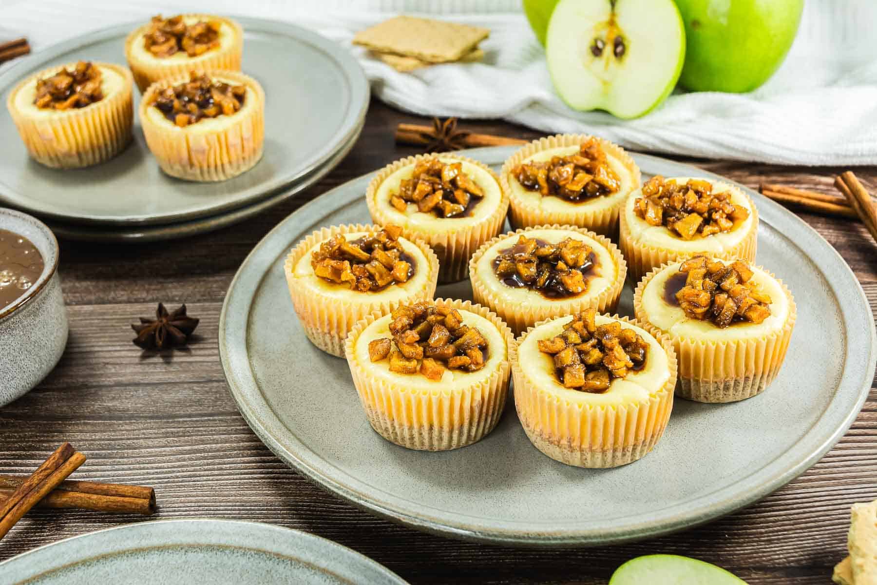 Mini cheesecakes topped with caramelized apple pieces on a gray plate, with green apples in the background.