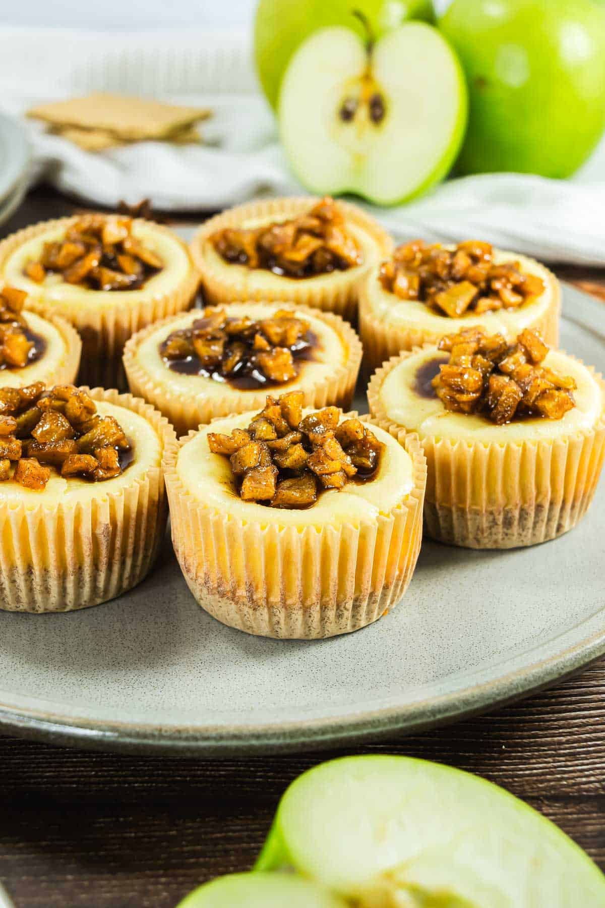 Mini cheesecakes topped with caramelized apple chunks on a gray plate, with green apples in the background.