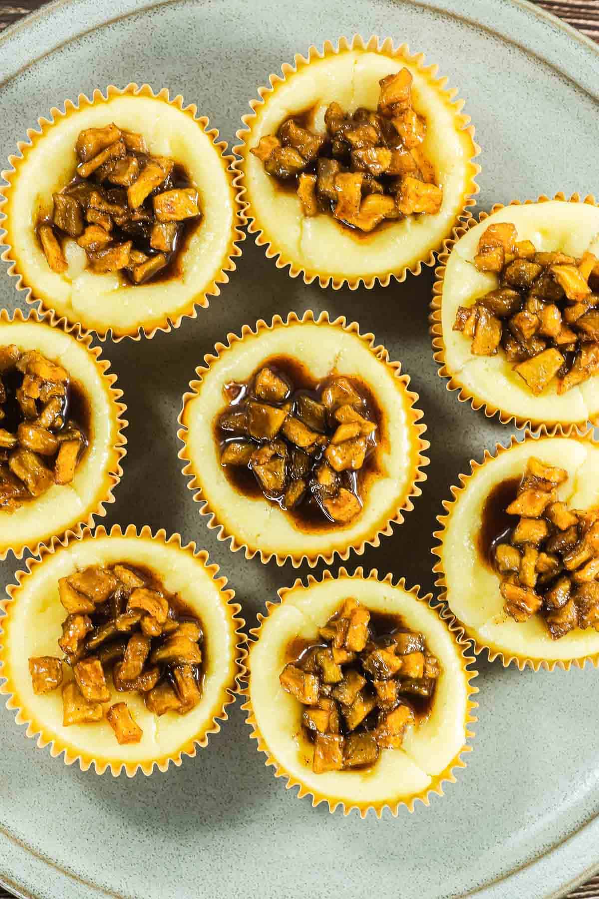 Mini cheesecakes topped with caramelized diced apples arranged in a circle on a gray plate.