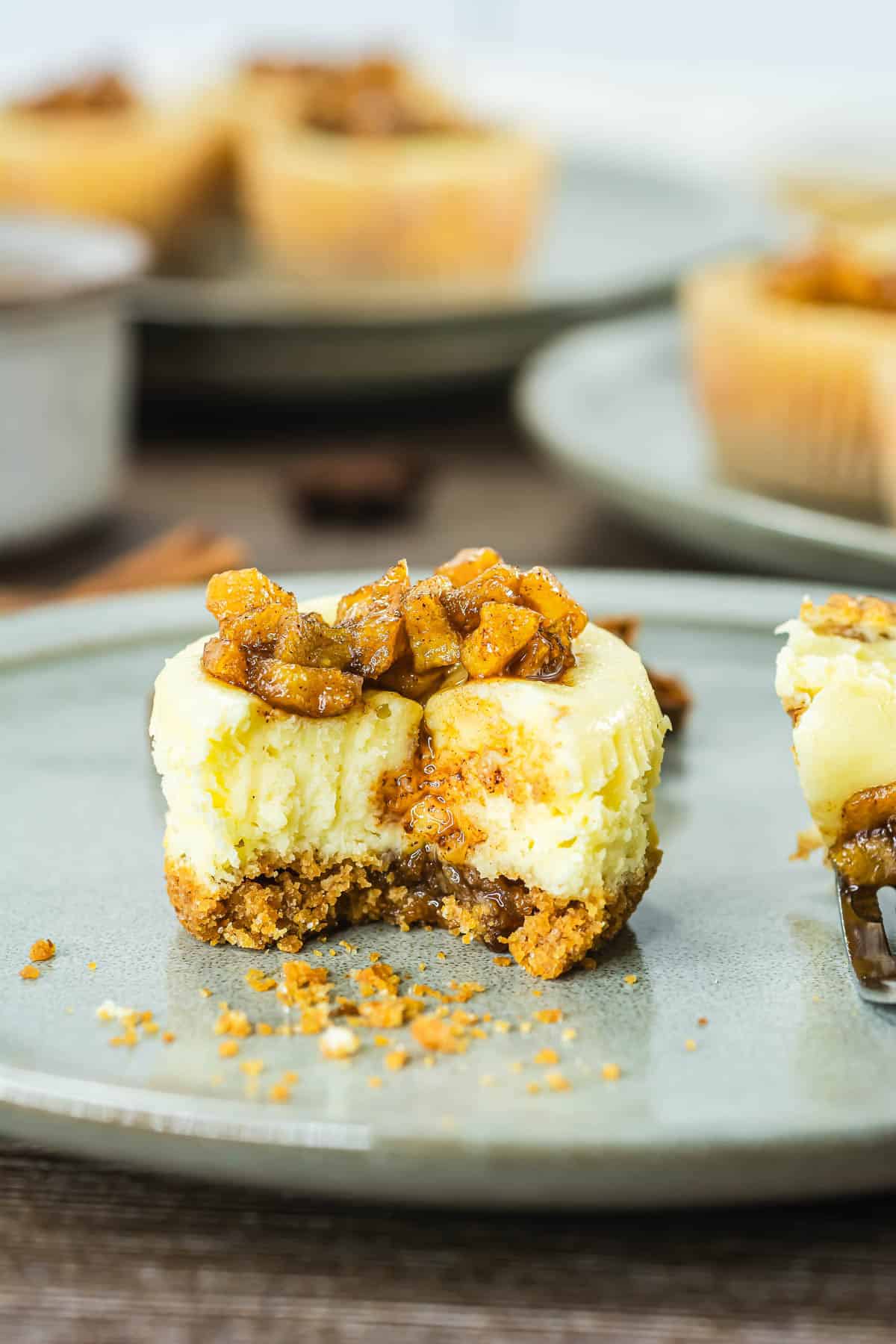 Mini cheesecake topped with spiced apple chunks, with a bite taken out, on a ceramic plate.