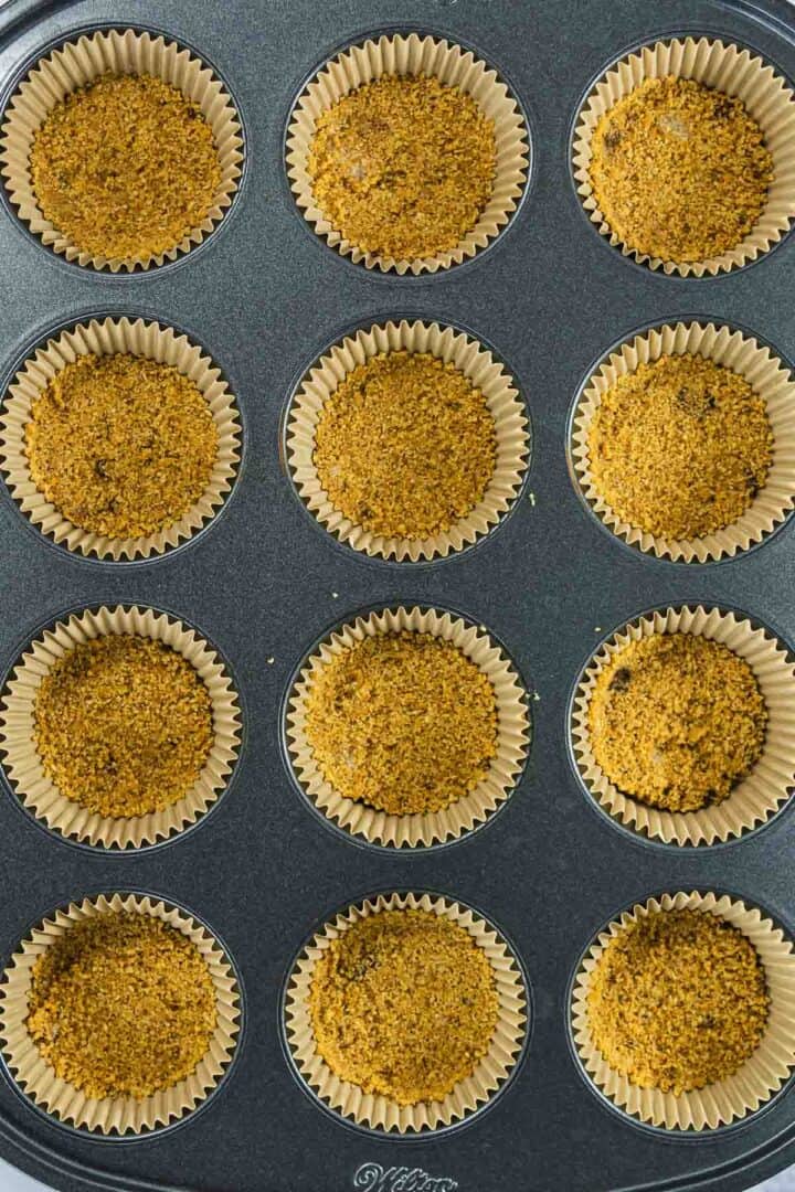 A muffin tin with 12 paper liners filled with crumbly graham cracker crusts.