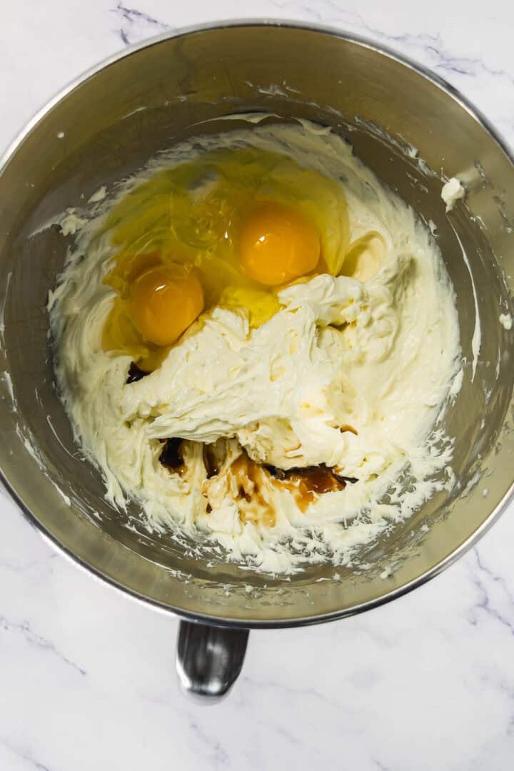 Stainless steel bowl with creamed butter, sugar, two eggs, and vanilla extract on a marble surface.