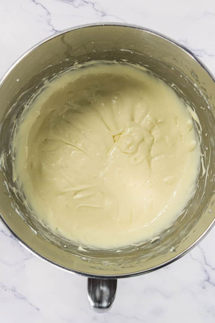 Creamy, pale yellow cheesecake batter in a metal mixing bowl on a white marble countertop.