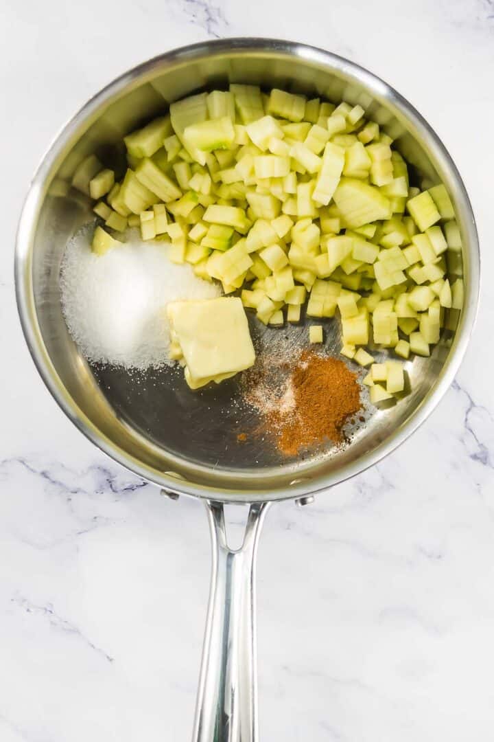 A saucepan with chopped apples, butter, sugar, and cinnamon on a marble surface.