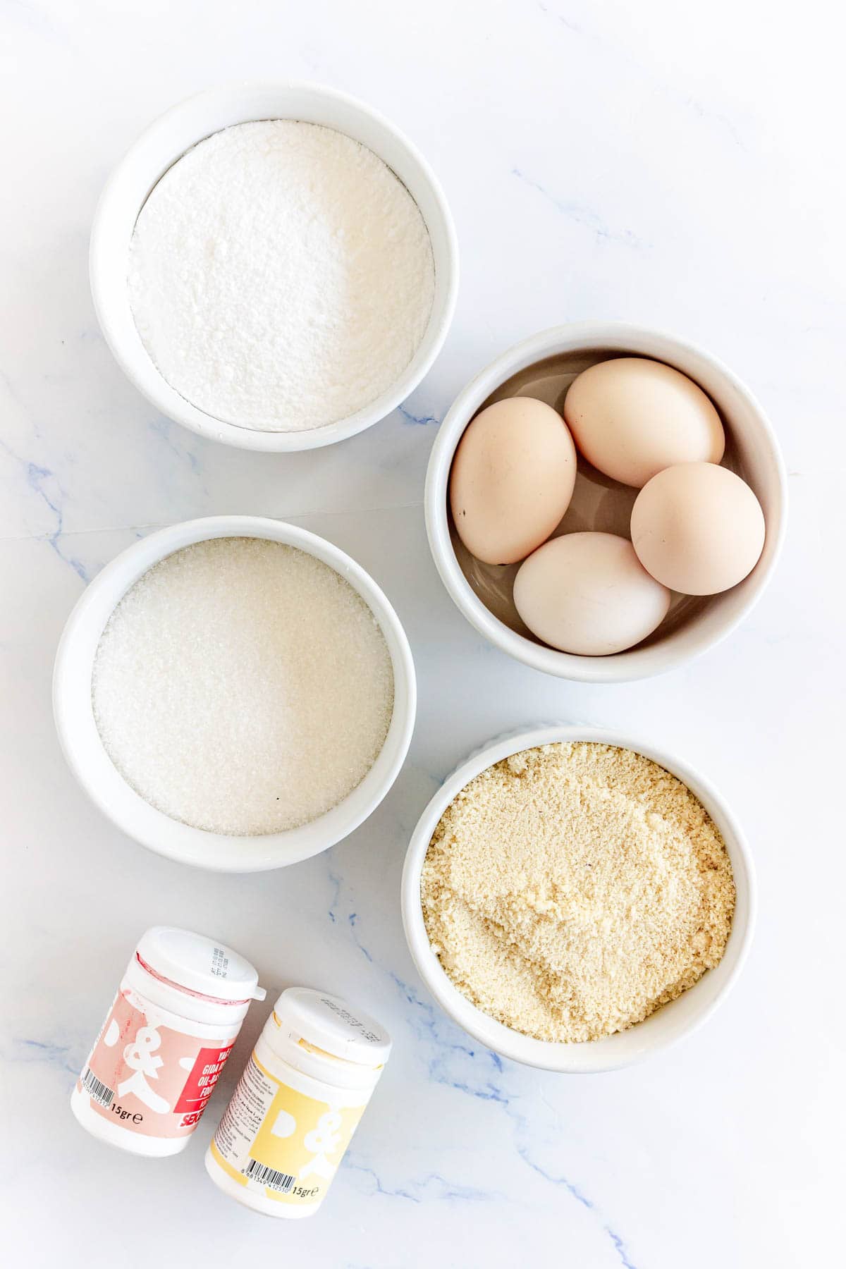 Five bowls with ingredients for Pumpkin Macarons: eggs, flour, sugar, almond flour, powdered sugar, and two small baking powder containers.