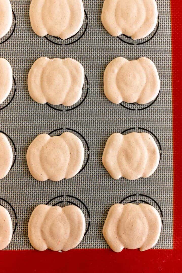 Pumpkin Macarons shells, shaped like mini pumpkins, rest neatly in rows on a baking mat, ready to be baked.