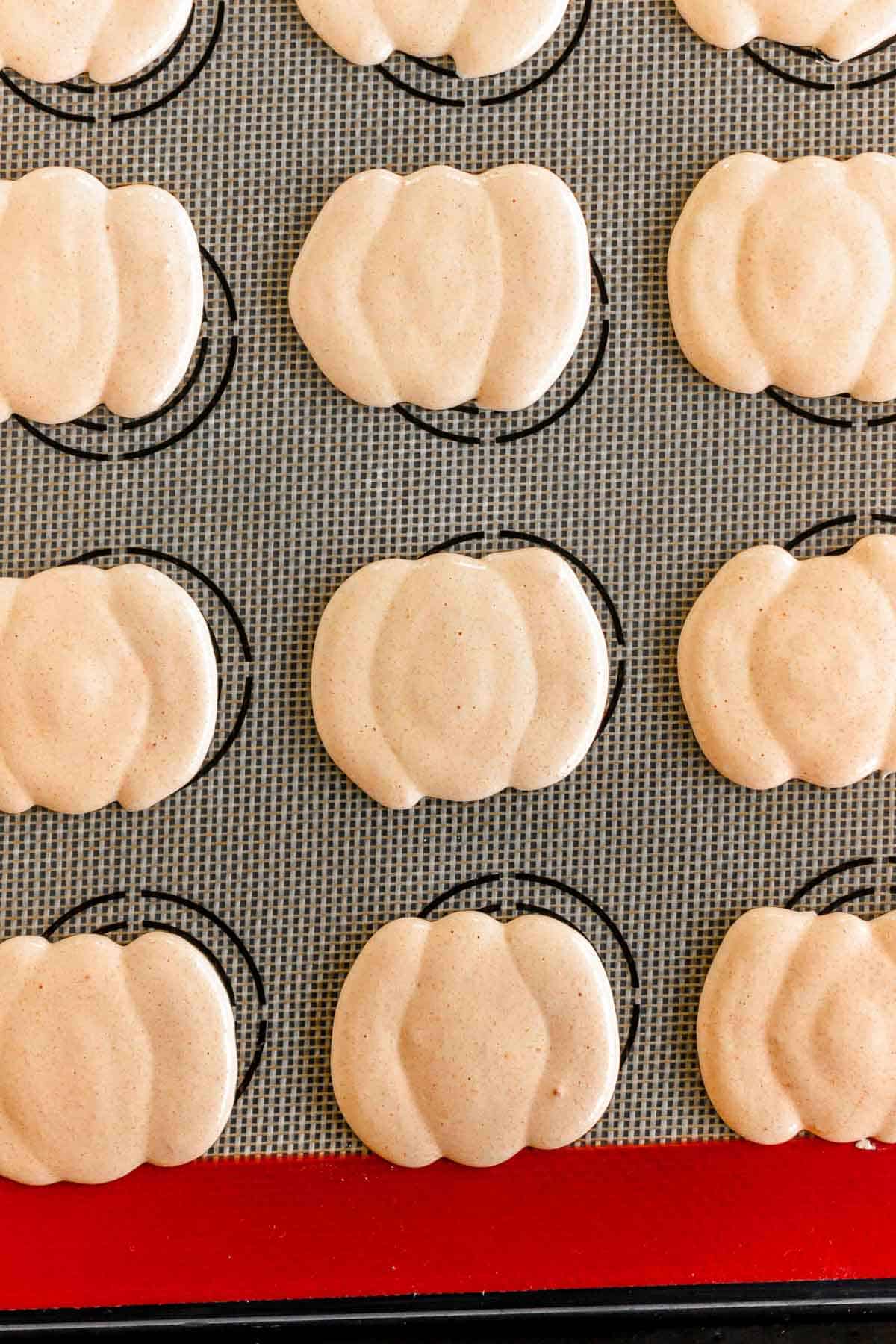 Pumpkin Macarons in festive pumpkin shapes rest on a baking mat with circular guides, ready for filling.