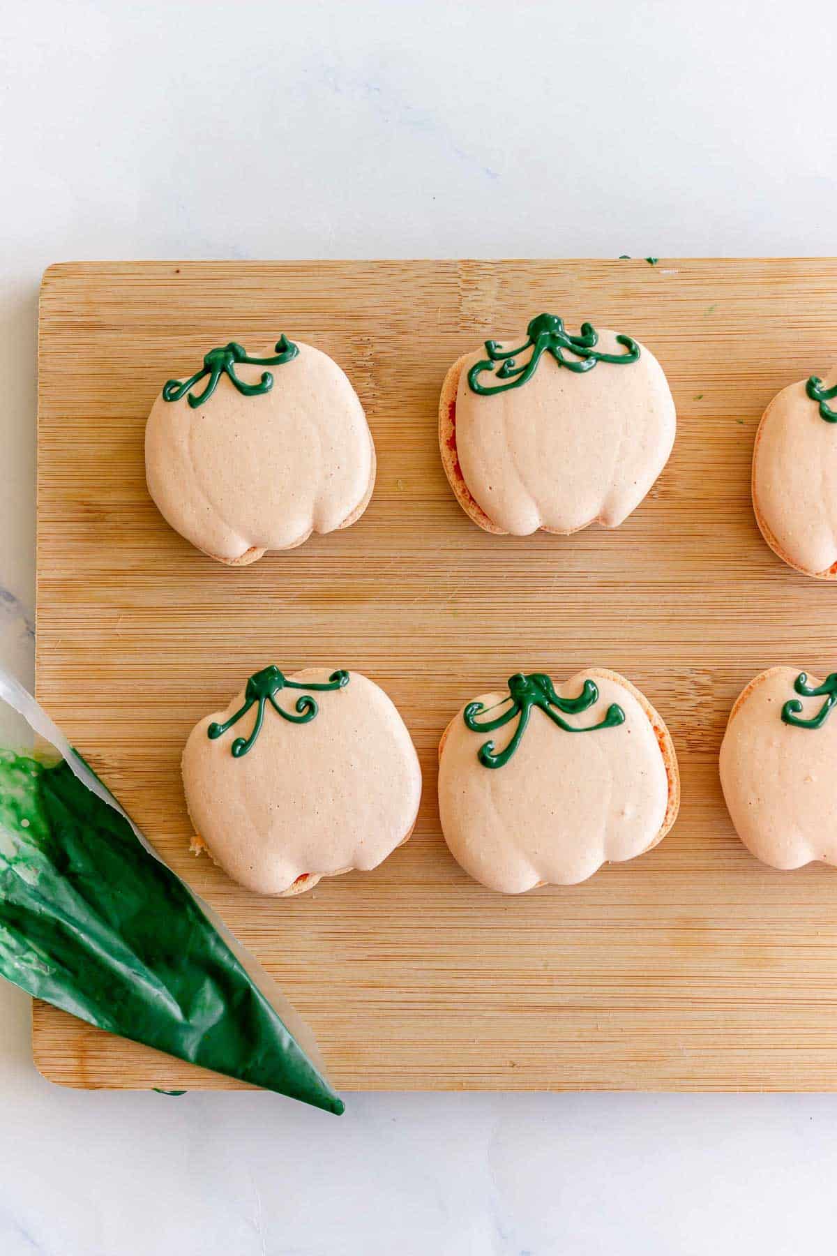 Pumpkin Macarons shaped like pumpkins with green icing stems rest on a wooden board, beside a green icing piping bag.