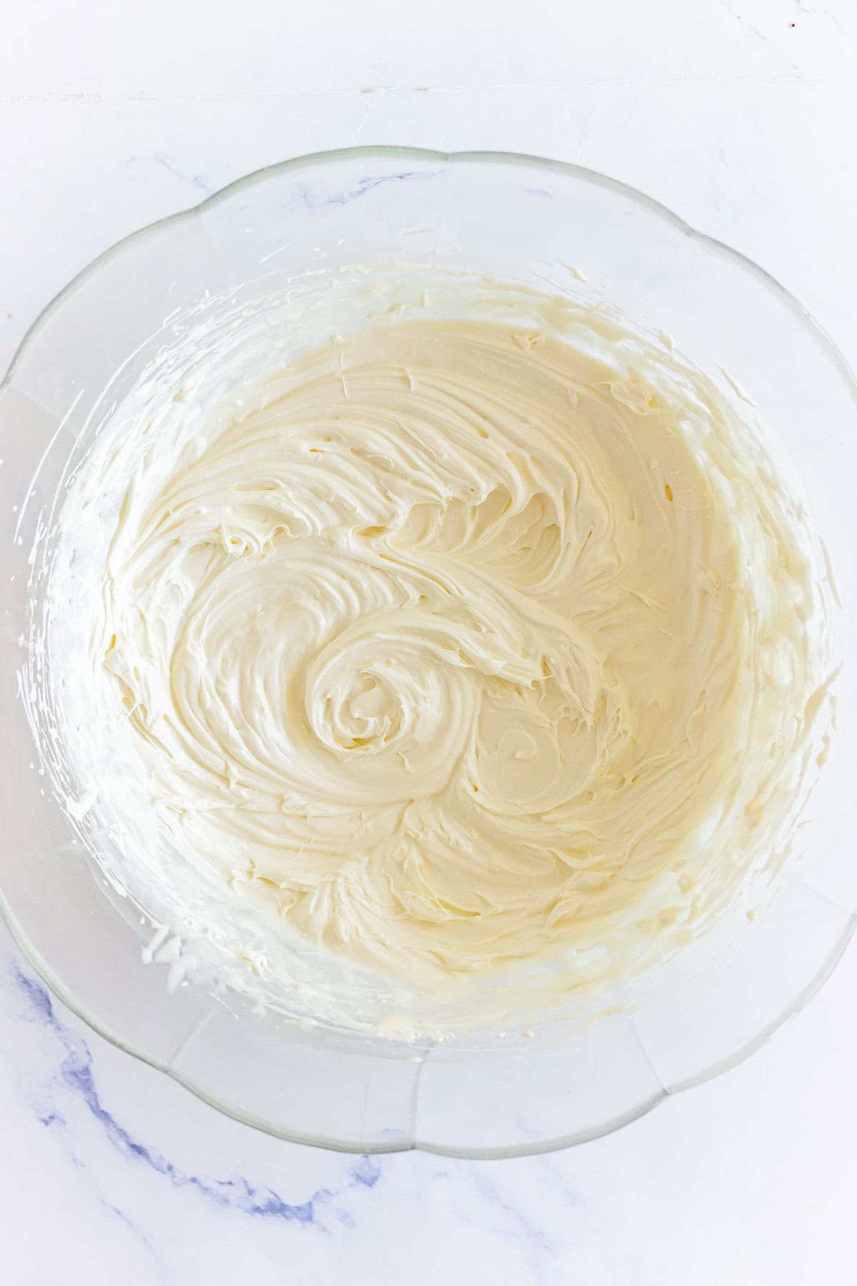 A glass bowl filled with smooth, swirled cream cheese frosting on a white surface, perfect for pairing with Pumpkin Macarons.