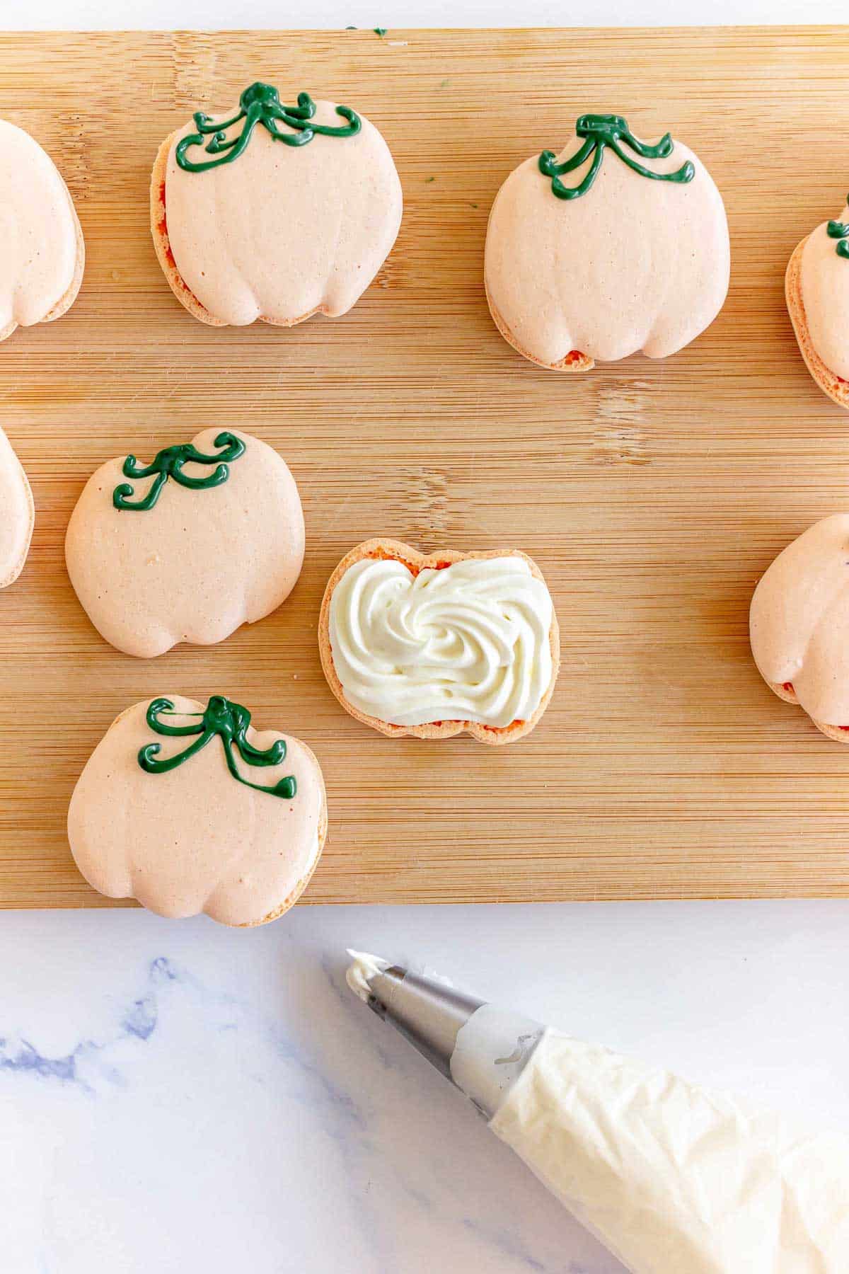 Pumpkin Macarons shaped like pumpkins rest on a wooden board, one open to reveal white filling, with an icing bag nearby.